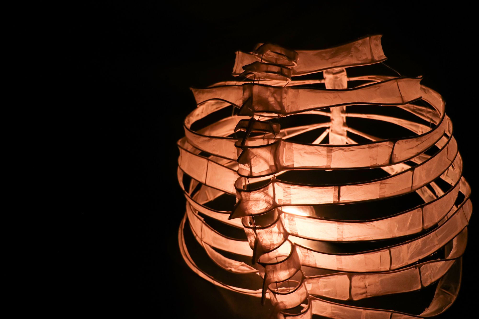 Close-up shot of the dress illuminated from within, emphasizing its glowing skeletal structure. The translucent material and curved bands create a ribcage-like form lit in warm amber tones.
